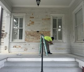a man removing worn paint on the outside of a historical home